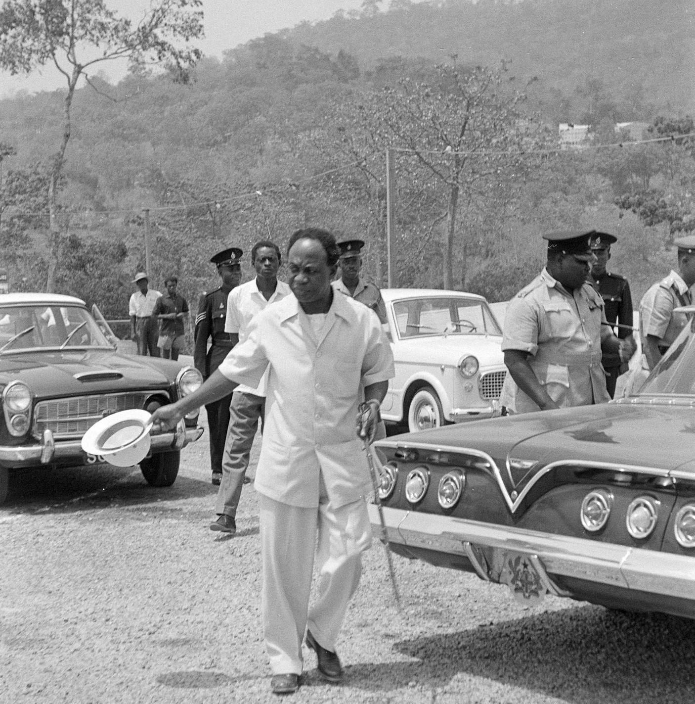 An African man in the foreground wearing a white suit and waving a white hat next to a 1960s Chevrolet car. More men, cars and forest in the background.